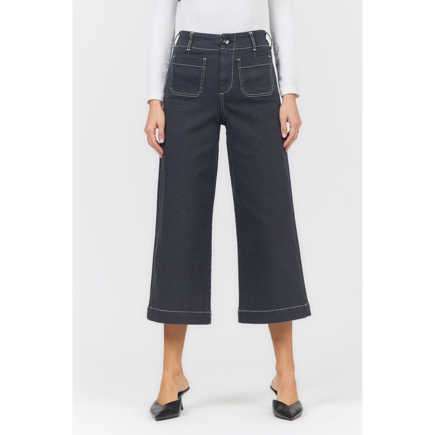A model wearing dark charcoal gray wide-leg cropped denim jeans with white contrast stitching, paired with a white long-sleeve top and black pointed-toe mule heels, shown against a plain light gray background.