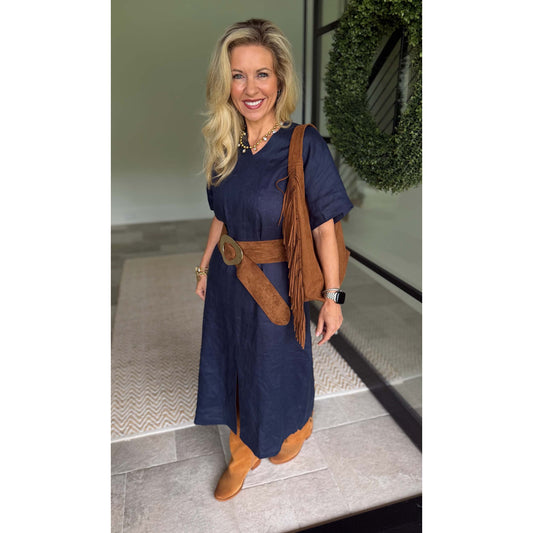A woman modeling a navy blue dress with a brown faux-suede belt, matching fringe detail, and tan boots, standing in a modern interior setting.