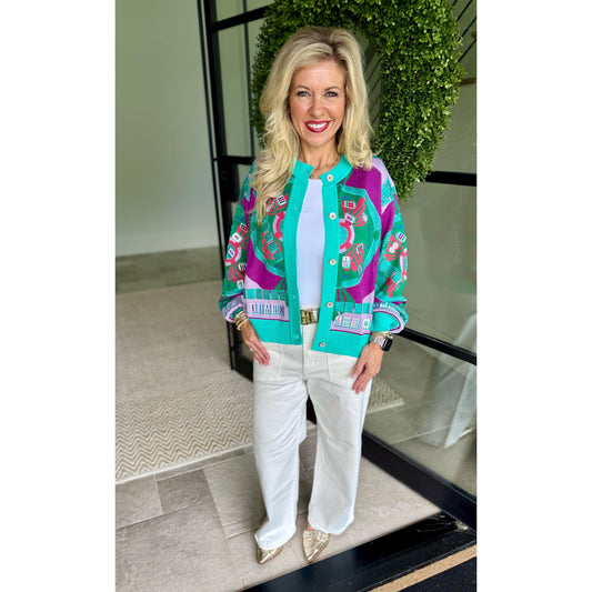 A woman stands in a doorway wearing a bright turquoise, magenta, and purple patterned button-up jacket over a white top and white pants, with gold pointed shoes.