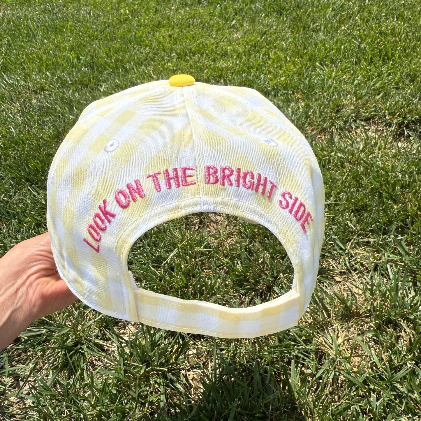 White cap with yellow polka dots and pink text 'Look on the Bright Side' held on grass