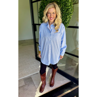 A woman wearing a light blue and white vertically striped button-down shirt, black leggings, and brown leather boots, standing in front of a glass door with a green wreath.