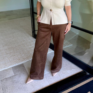 A person wearing a cream short-sleeve peplum-style top with a gold button detail and wide-leg brown pants, standing indoors on a tiled floor near a glass door.