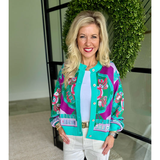 A smiling blonde woman stands indoors wearing a bright turquoise and magenta patterned bomber-style jacket over a white top and white pants. A green leafy wreath and glass doors are visible behind her.