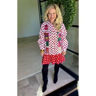 A woman stands indoors wearing a white long-sleeve blouse with red polka dots and colorful nutcracker appliques, paired with a red and white polka dot skirt, black tights, and black knee-high boots.