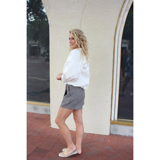 A woman stands outdoors in front of a cream-colored building, wearing a white long-sleeve blouse, a black and white gingham mini skirt, and clear espadrille flats.