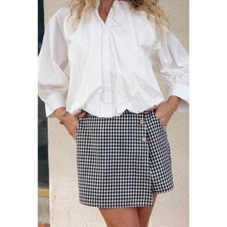 A woman wearing a white long-sleeve button-up blouse and a black-and-white gingham mini skirt with asymmetrical buttons, standing against a light beige wall.