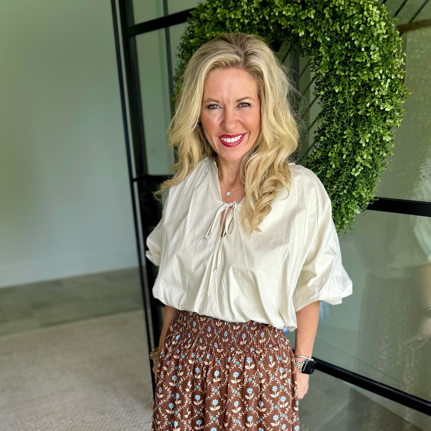 A smiling blonde woman wearing a cream-colored blouse with puff sleeves and a brown patterned skirt, standing in front of a glass door with a green leafy wreath.