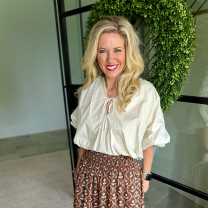 A smiling blonde woman wearing a cream-colored blouse with puff sleeves and a brown patterned skirt, standing in front of a glass door with a green leafy wreath.