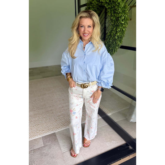 A woman stands indoors wearing a light blue button-up shirt with voluminous sleeves, white wide-leg pants with a pink floral print, and a gold belt.