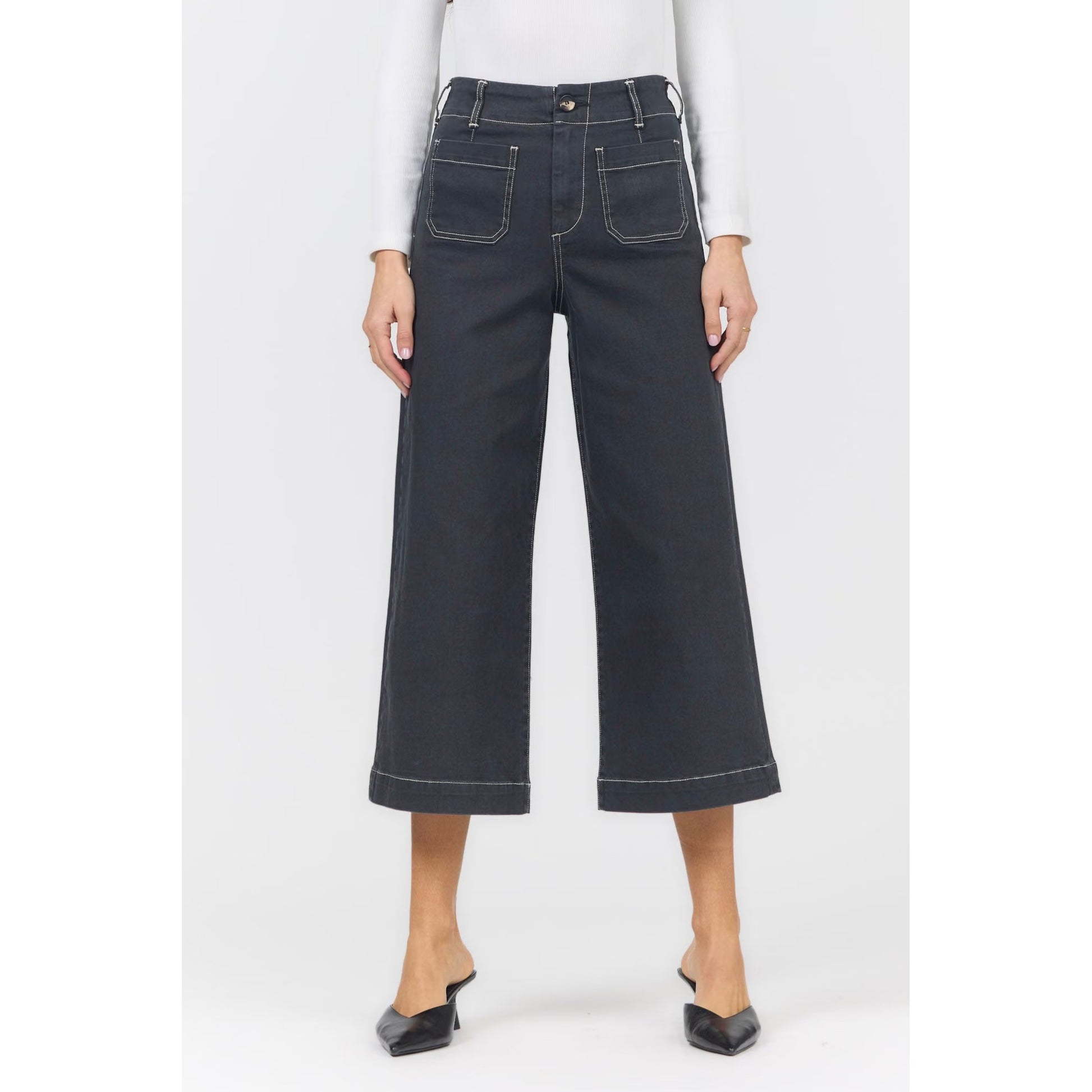 A model wearing dark charcoal gray wide-leg cropped denim jeans with white contrast stitching, paired with a white long-sleeve top and black pointed-toe mule heels, shown against a plain light gray background.