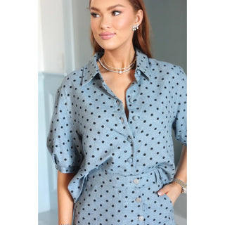 A woman wearing a light blue short-sleeve button-up shirt with black polka dots, styled with layered necklaces and earrings.