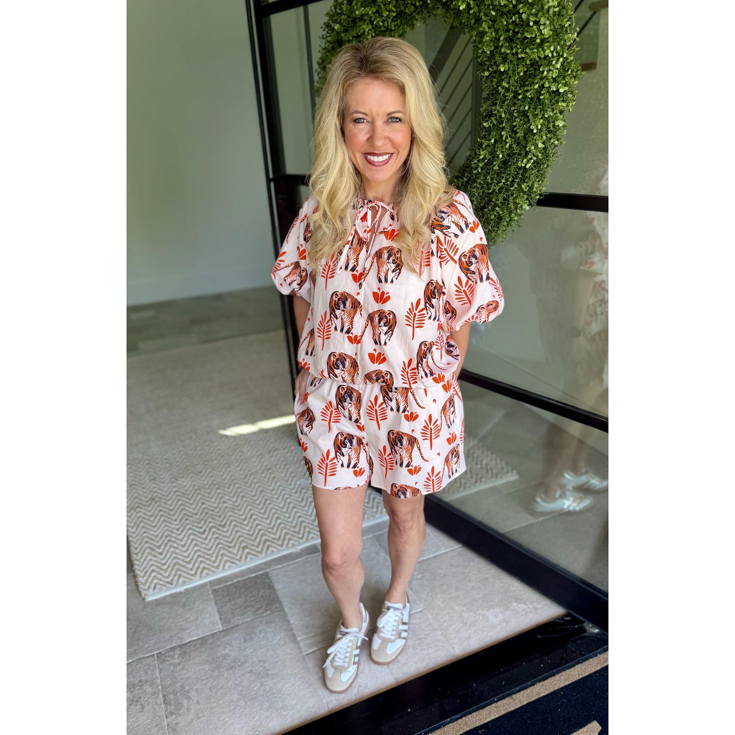 A woman stands in a doorway wearing a short, loose-fitting romper with a white base and an allover print of brown dogs and orange leaves. She is wearing white sneakers, with a green wreath and glass door visible in the background.