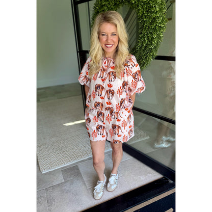 A woman stands in a doorway wearing a short, loose-fitting romper with a white base and an allover print of brown dogs and orange leaves. She is wearing white sneakers, with a green wreath and glass door visible in the background.