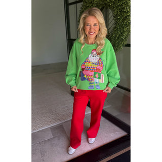 A woman modeling a bright green crewneck sweatshirt with a colorful Santa Claus graphic, paired with red pants and white sneakers, standing on a tiled floor in front of a glass door.