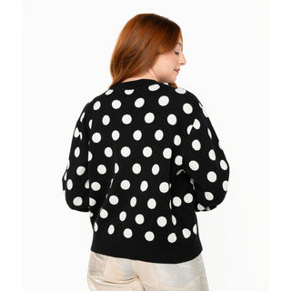 A woman with red hair is shown from the back wearing a black knit sweater with a white polka dot pattern, paired with light beige pants, against a plain white background.