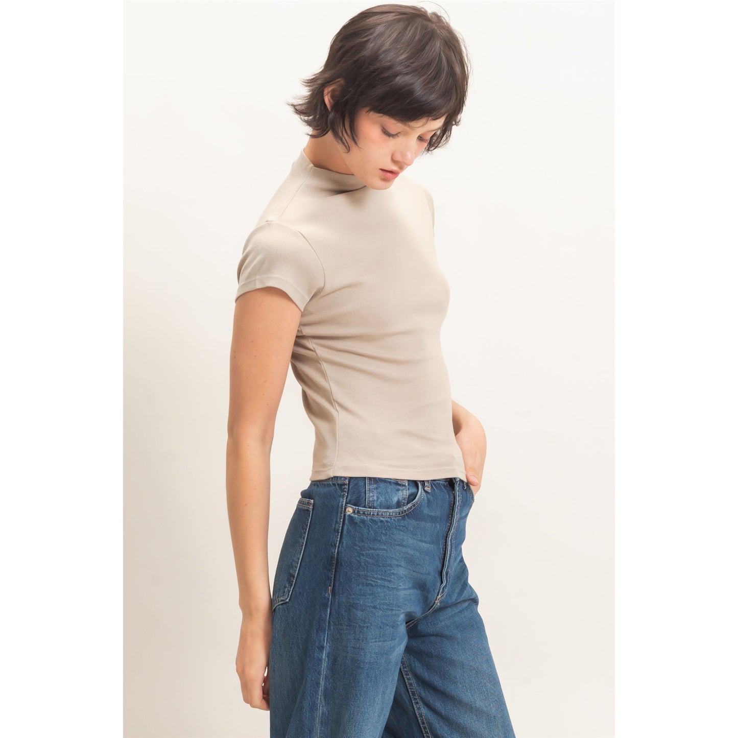 A woman wearing a beige short-sleeve mock neck t-shirt and blue jeans, standing against a plain off-white background.