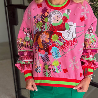 A pink Christmas-themed crewneck sweatshirt with a colorful embroidered design of Santa in a sleigh, worn by a model in green pants.