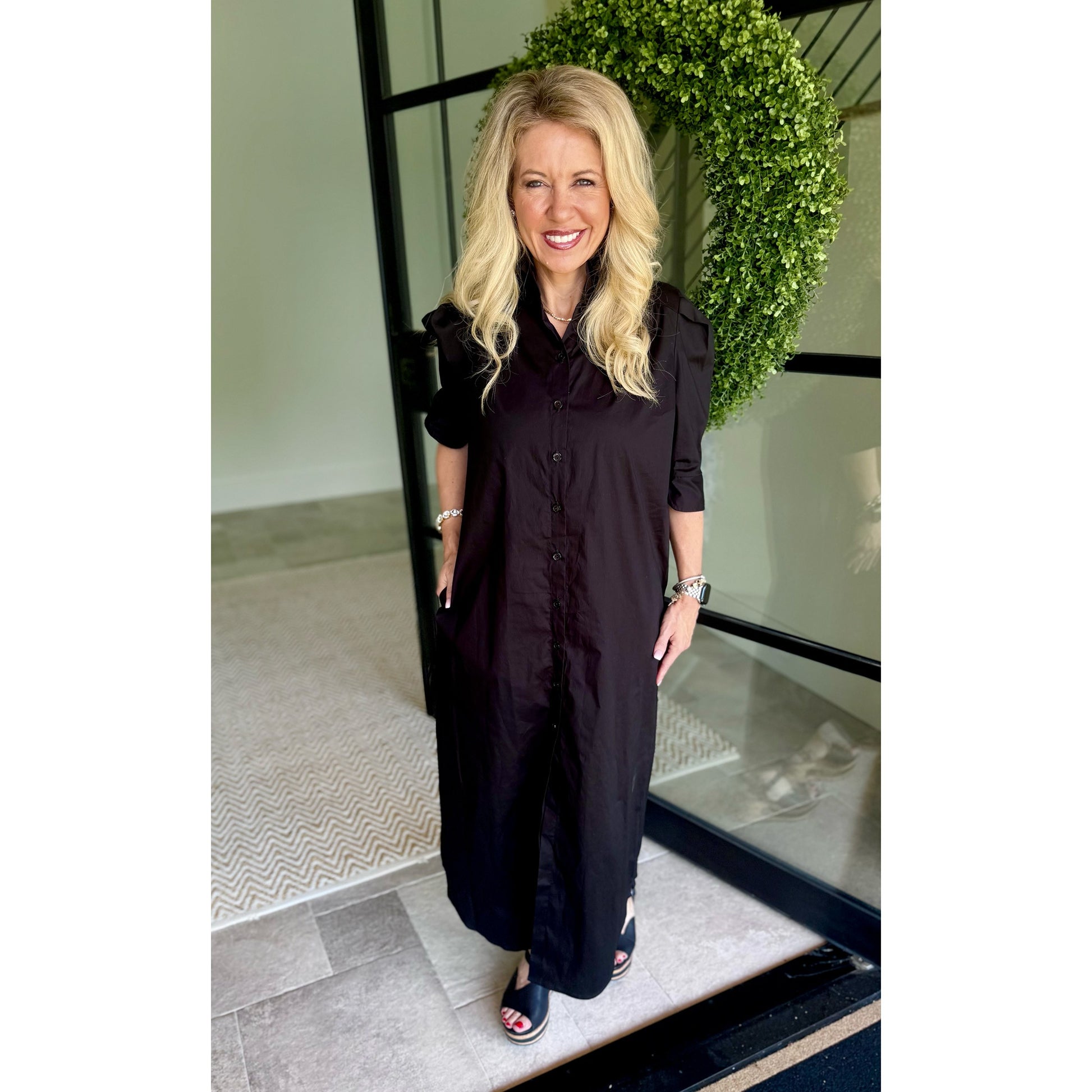 A woman with blonde hair wearing a long black button-up shirt dress, standing in front of a glass door with a green wreath in the background.