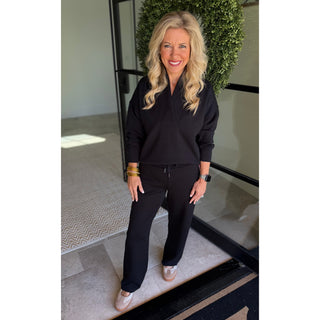 A woman with blonde hair stands in front of a glass door wearing a matching black long-sleeve top and wide-leg pants, with gold bracelets and white sneakers.
