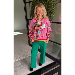 A woman wearing a vibrant pink Christmas-themed crewneck sweater with colorful holiday graphics and green wide-leg trousers, standing in front of a glass door and green wreath.