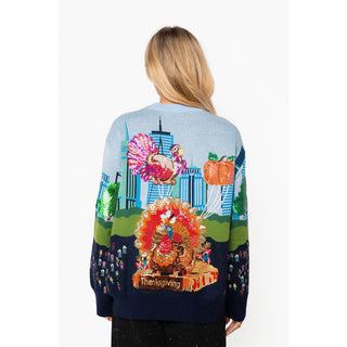 A woman viewed from behind wearing a light blue and navy knit sweater with a large Thanksgiving-themed graphic on the back, featuring turkeys, pumpkins, a cornucopia, and a city skyline.