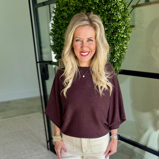 A smiling blonde woman wearing a loose, short-sleeve scoop-neck top in deep plum-burgundy and cream pants, standing indoors in front of a green hedge and glass railing.