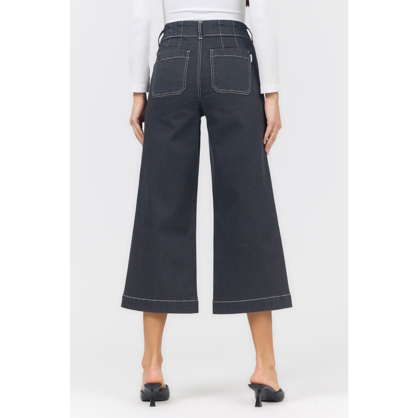 Back view of a model wearing dark charcoal wide-leg cropped jeans with white contrast stitching, paired with a white long-sleeve top and black mule heels, on a light gray studio background.