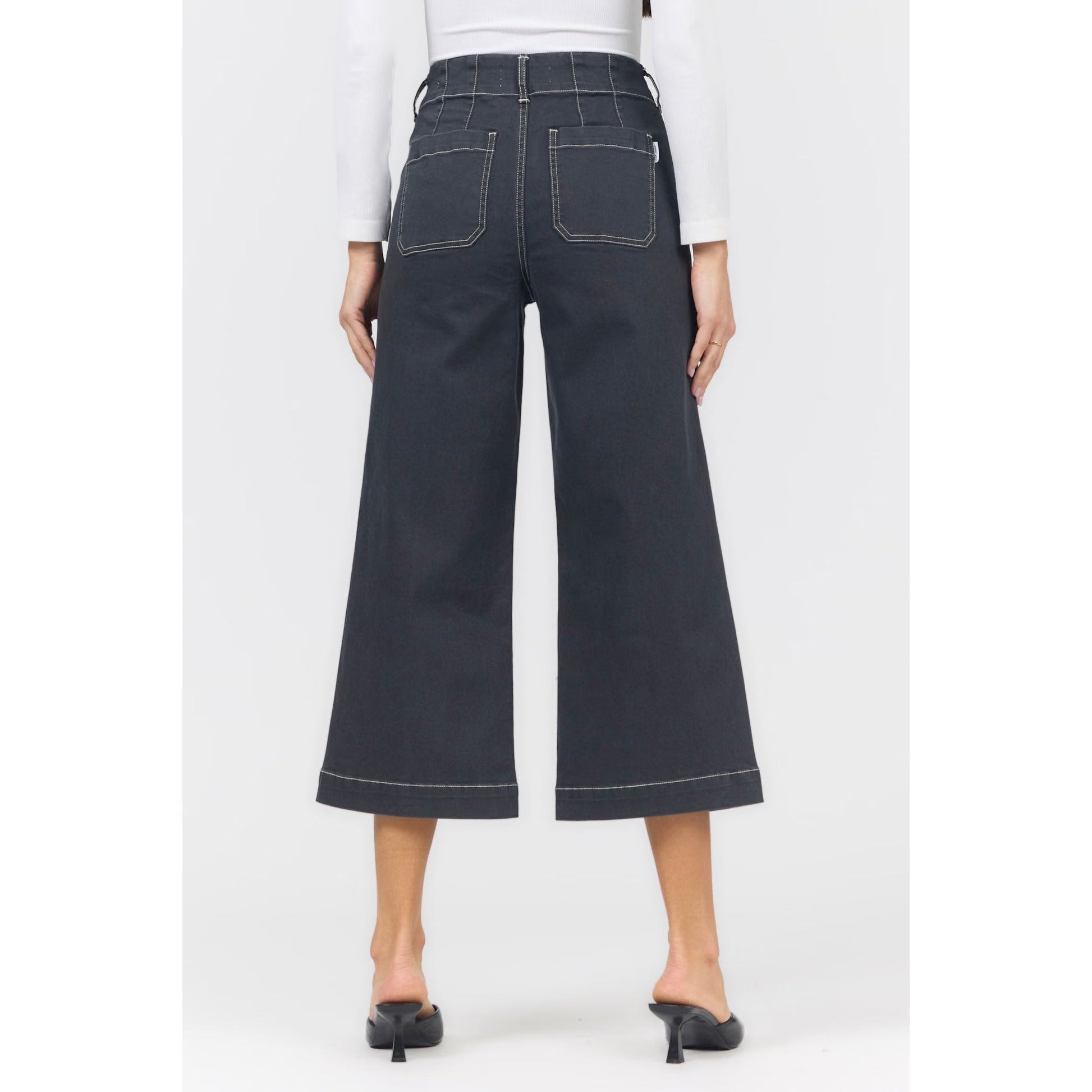 Back view of a model wearing dark charcoal wide-leg cropped jeans with white contrast stitching, paired with a white long-sleeve top and black mule heels, on a light gray studio background.