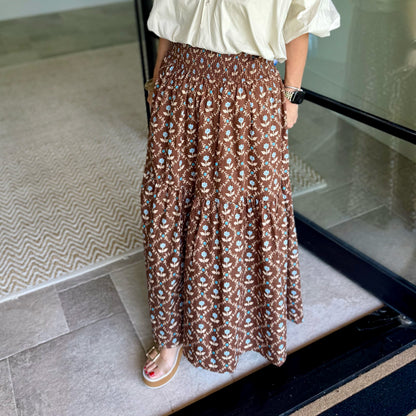 A person wearing a brown smocked maxi skirt with a white and blue floral pattern, paired with a cream blouse and tan sandals, standing on a tiled floor next to a glass door.