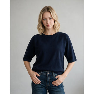 A woman with blonde hair wearing a dark navy blue short-sleeve knit sweater and blue jeans, standing against a neutral gray studio background.