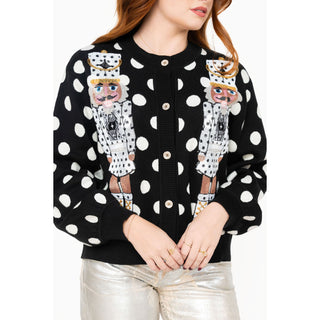 A woman wearing a black cardigan sweater with white polka dots and two large nutcracker figures on the front, paired with metallic silver pants, against a plain white background.