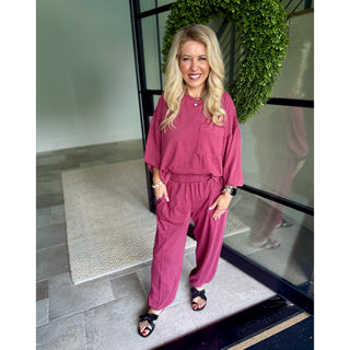 A woman stands indoors wearing a matching dusty rose two-piece lounge set with a short-sleeve top and wide-leg pants, paired with black slide sandals. She is positioned in front of a glass door with a green wreath on the frame.