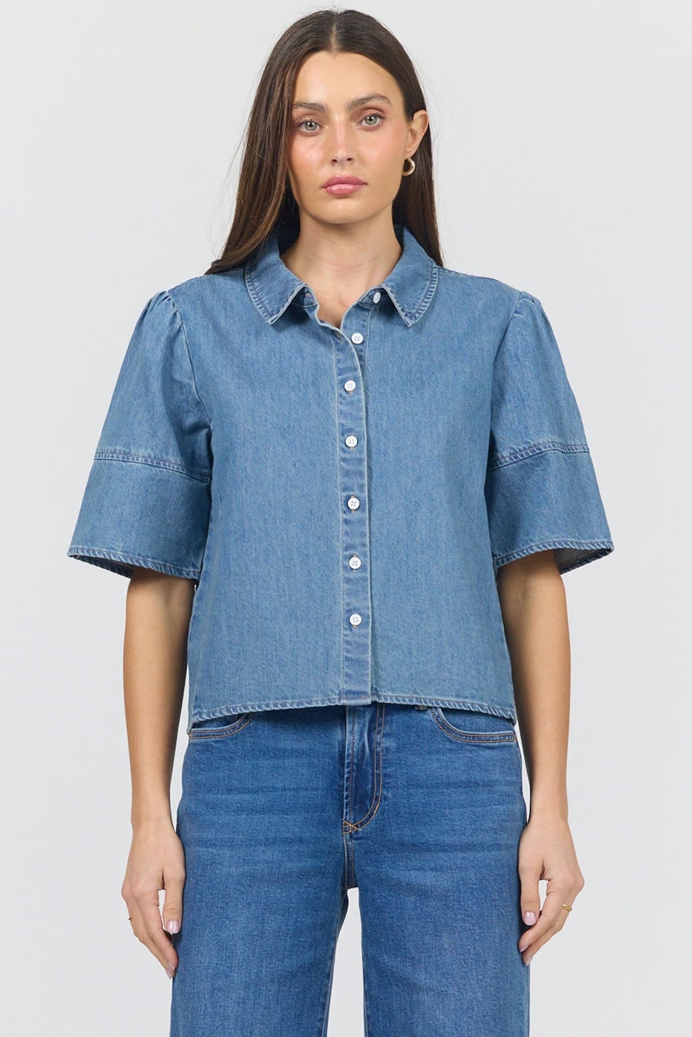 A woman modeling a cropped blue denim short-sleeve button-up shirt with puffed sleeves, paired with blue jeans, against a light gray studio background.