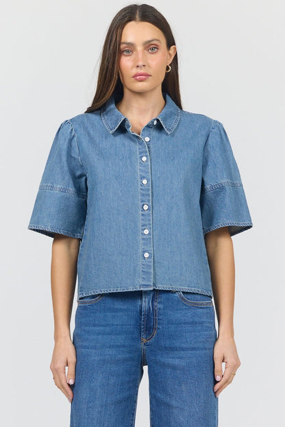 A woman modeling a cropped blue denim short-sleeve button-up shirt with puffed sleeves, paired with blue jeans, against a light gray studio background.