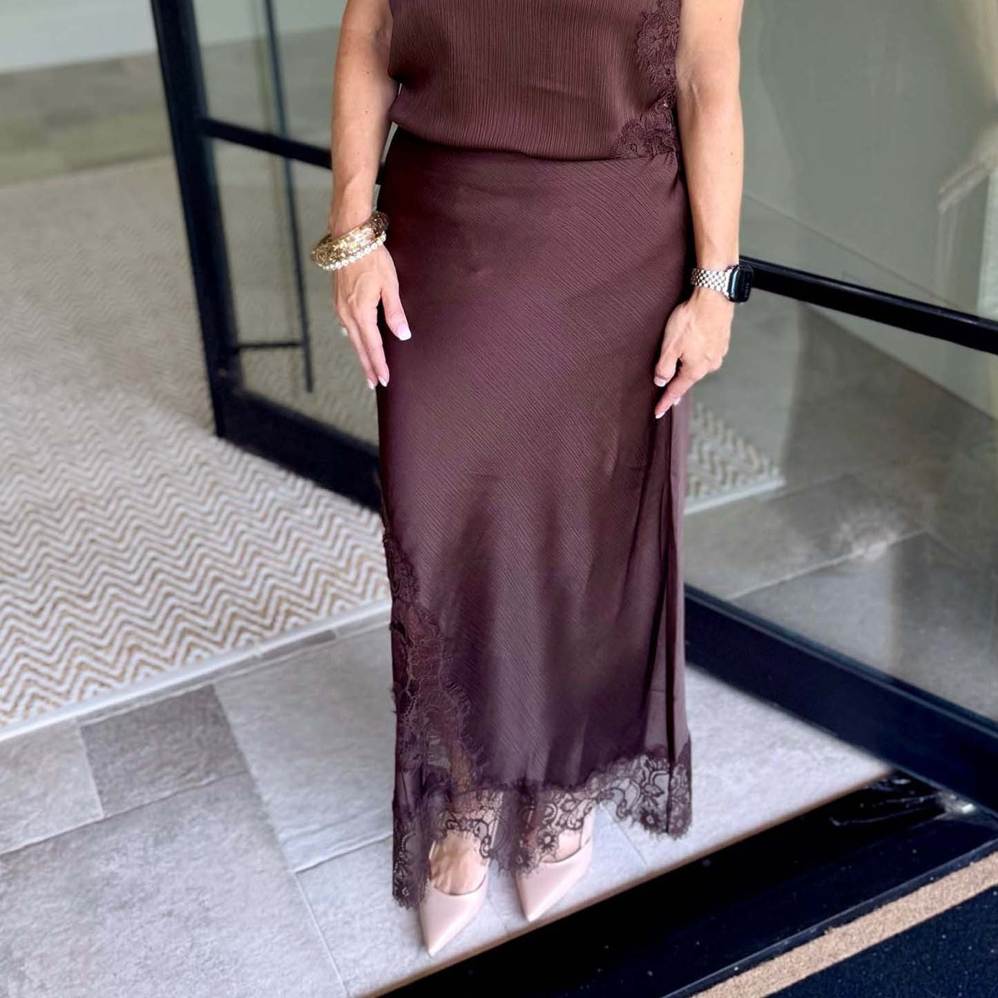 A woman wearing a dark brown ribbed midi skirt with lace trim at the hem, paired with a matching top and nude pointed-toe heels, standing on a tiled entryway.