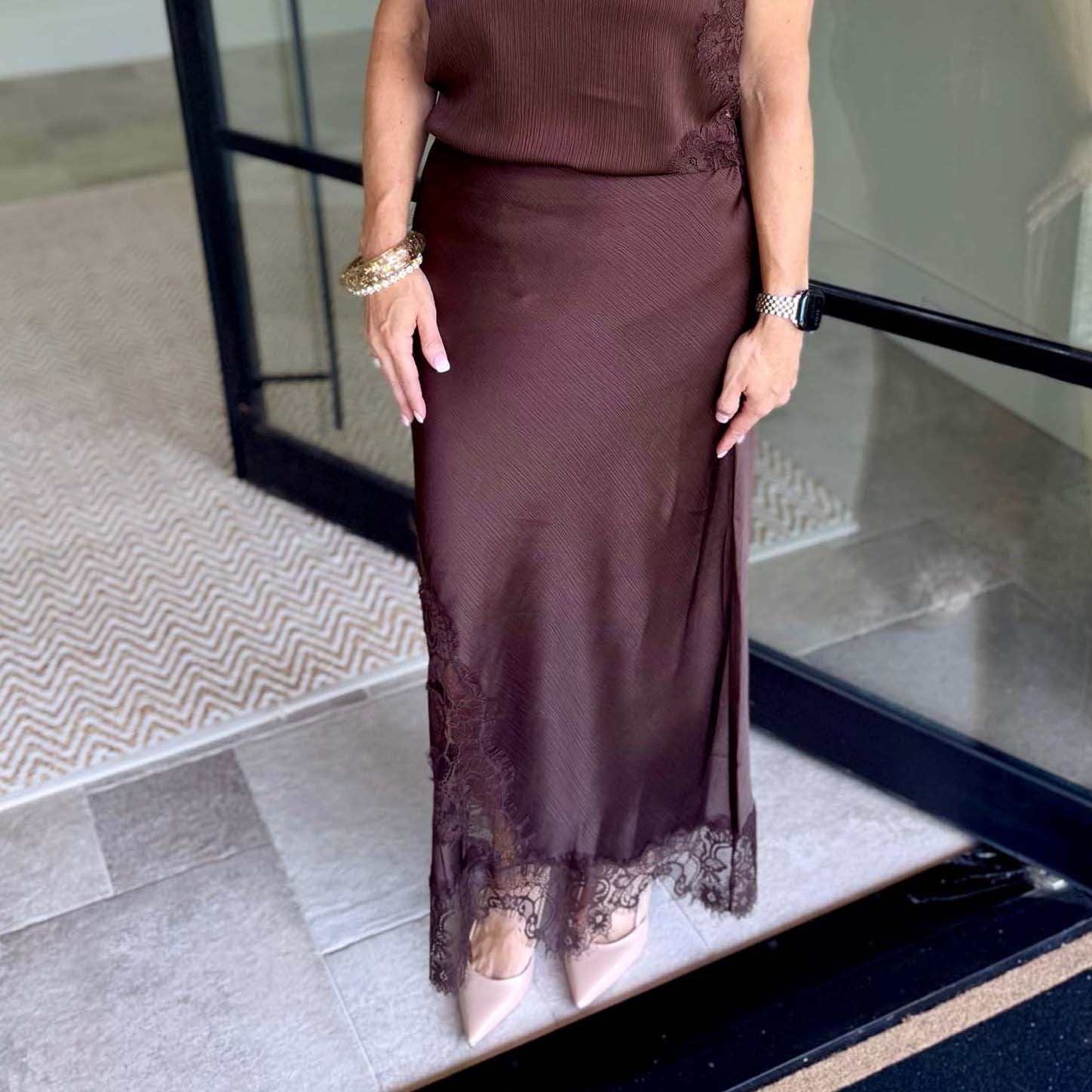 A woman wearing a dark brown ribbed midi skirt with lace trim at the hem, paired with a matching top and nude pointed-toe heels, standing on a tiled entryway.