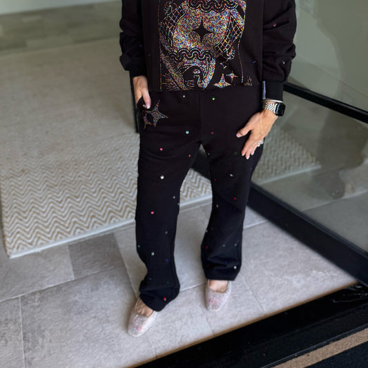 A person wearing a black embellished outfit with a star-patterned top and black pants decorated with small colorful rhinestones, standing on a tiled floor.