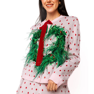 A woman wearing a light pink knit sweater with red polka dots, a red velvet bow, and a green artificial wreath with a red ribbon, paired with a matching skirt against a white background.