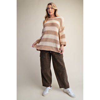 A woman modeling an oversized knit sweater with horizontal beige and cream stripes, paired with brown wide-leg patterned pants and white sneakers, against a plain beige studio background.