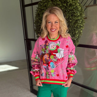 A smiling blonde woman wearing a bright pink Christmas-themed crewneck sweater with a festive Santa graphic and green pants, standing indoors in front of a glass door and green wreath.