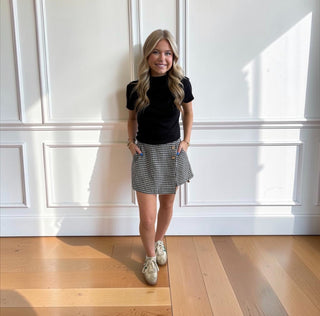 A woman stands in a bright room with white paneled walls and light wood floors, wearing a black short-sleeve mock-neck top, a black-and-white gingham mini skirt, and white sneakers.
