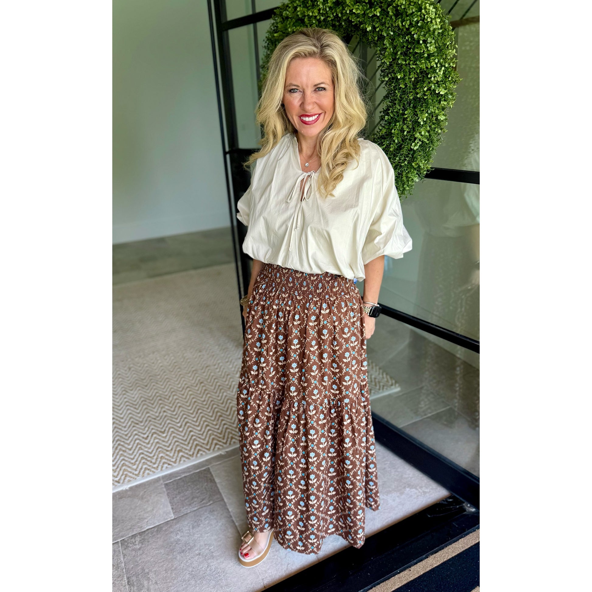 A woman stands indoors wearing a cream-colored blouse with puff sleeves and a brown maxi skirt featuring a white and beige geometric pattern. She is posing in front of a glass door with a green wreath in the background.