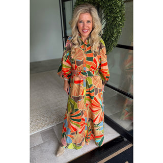 A smiling blonde woman stands indoors wearing a long-sleeve maxi dress with a bold retro fan and leaf pattern in orange, green, beige, and blue. The dress has a tie waist and is photographed in front of a glass door with a green wreath in the background.
