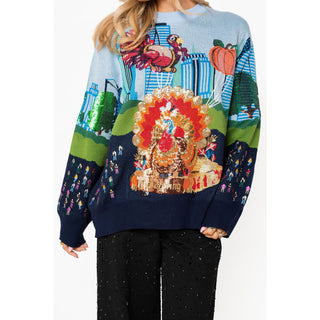 A woman wearing a long-sleeve knit sweater with a colorful Thanksgiving-themed graphic featuring a large turkey, pumpkins, skyscrapers, and small figures, paired with black pants against a plain white background.