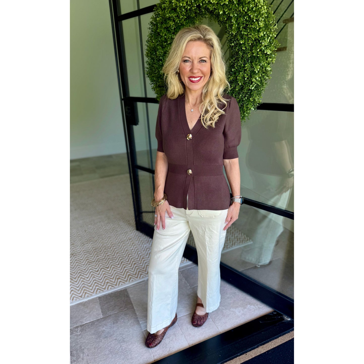 A woman stands in front of a glass door with a green wreath, wearing a short-sleeve brown button-front top with gold buttons and white wide-leg pants.
