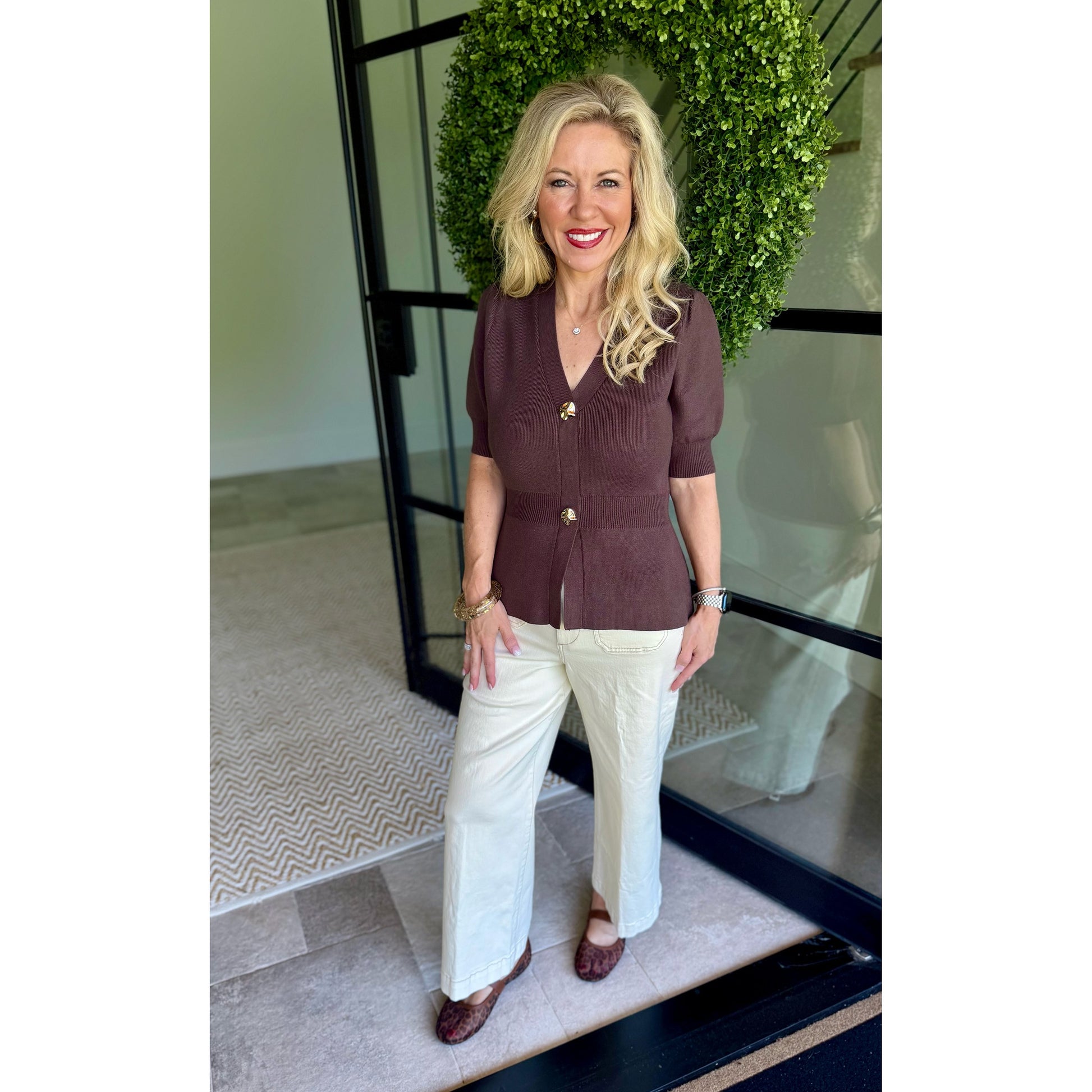 A woman stands in front of a glass door with a green wreath, wearing a short-sleeve brown button-front top with gold buttons and white wide-leg pants.