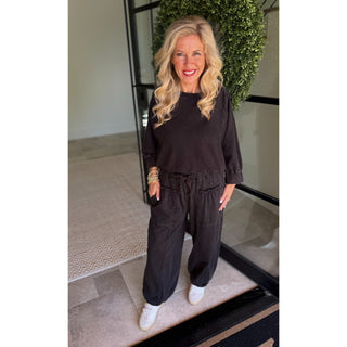 A woman modeling a dark brown lounge set consisting of a long-sleeve crewneck top and matching drawstring pants, paired with white sneakers, standing in a modern entryway.