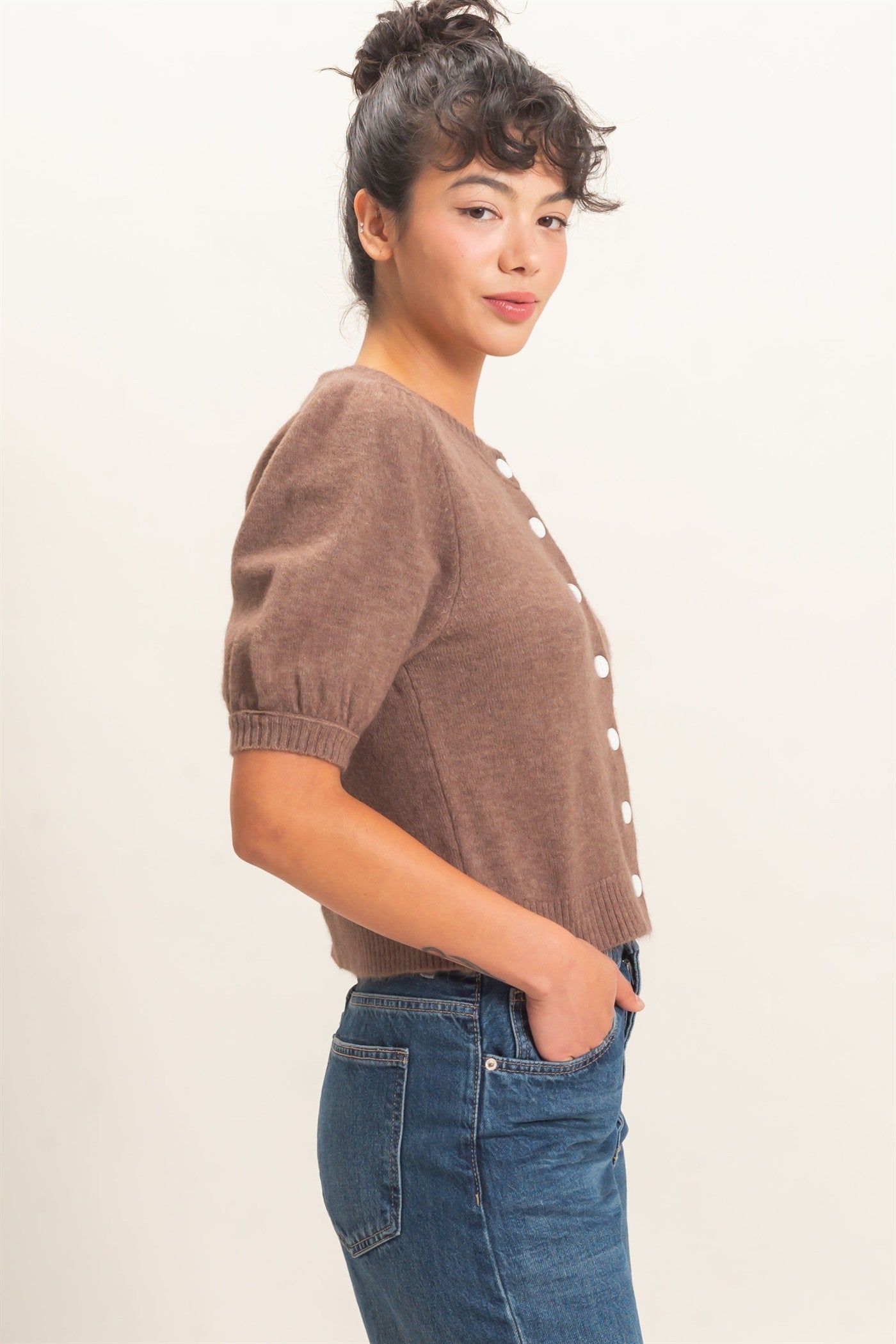 Woman wearing a brown sweater with white buttons and blue jeans on a plain background