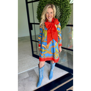 A woman stands in front of a glass door wearing a long-sleeve dress with a bold blue, yellow, and red abstract print, a large red bow at the neckline, and matching blue cowboy boots.