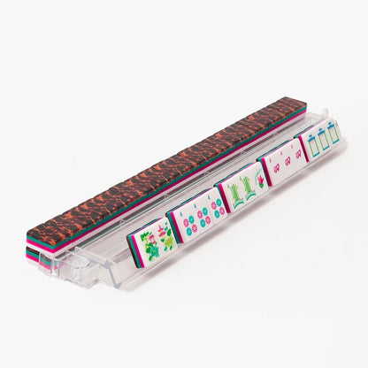 A clear acrylic mahjong rack holding multiple colorful mahjong tiles, with a textured dark brown and black patterned tile resting on top, photographed on a clean white background.